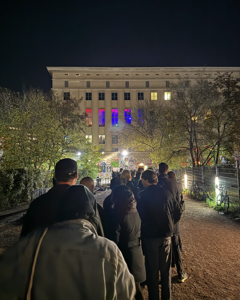 A picture taken from the line to get in to Berghain. The building is in the background. It is night time and you can see a long line up of people.