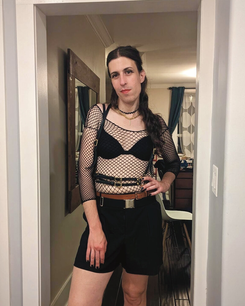 A picture of me. I have eyeliner, eyeshadow, and lipstick on, and I am wearing a golden necklace with black mesh top over a black bra with a black leather harness, a brown belt, and black shorts.