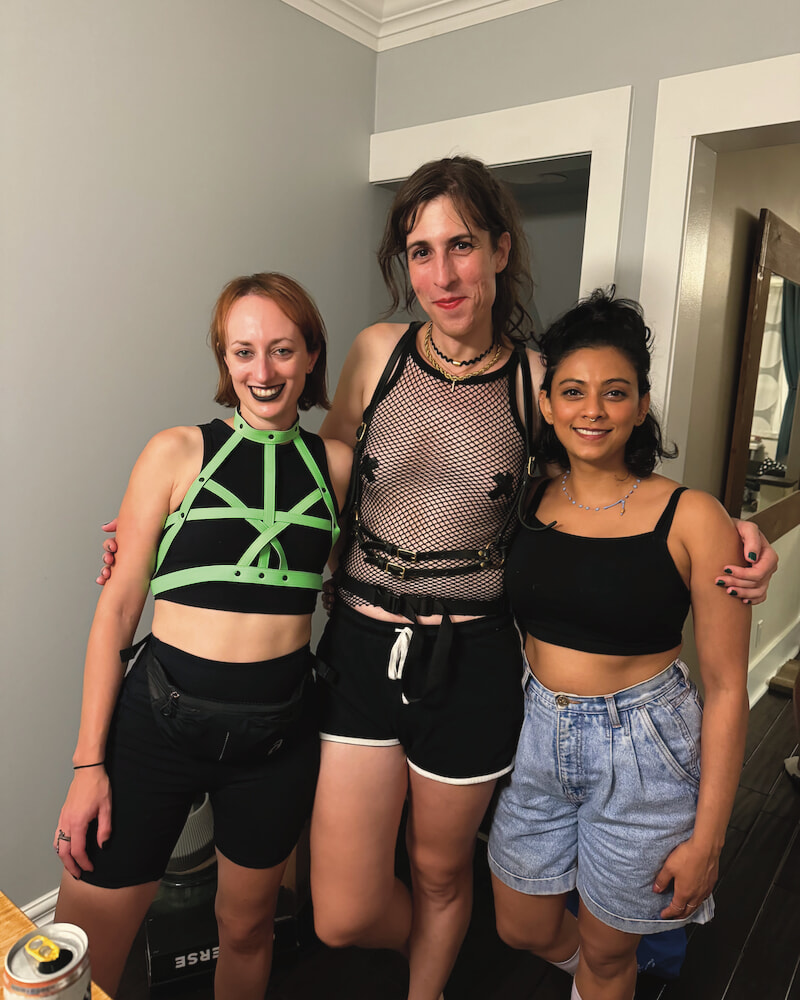 Me and two friends are standing together, my arms around them. We're all dressed to go out dancing. I'm wearing a mesh tank top, short shorts, a leather harness and my nipples are covered with electrical tape.