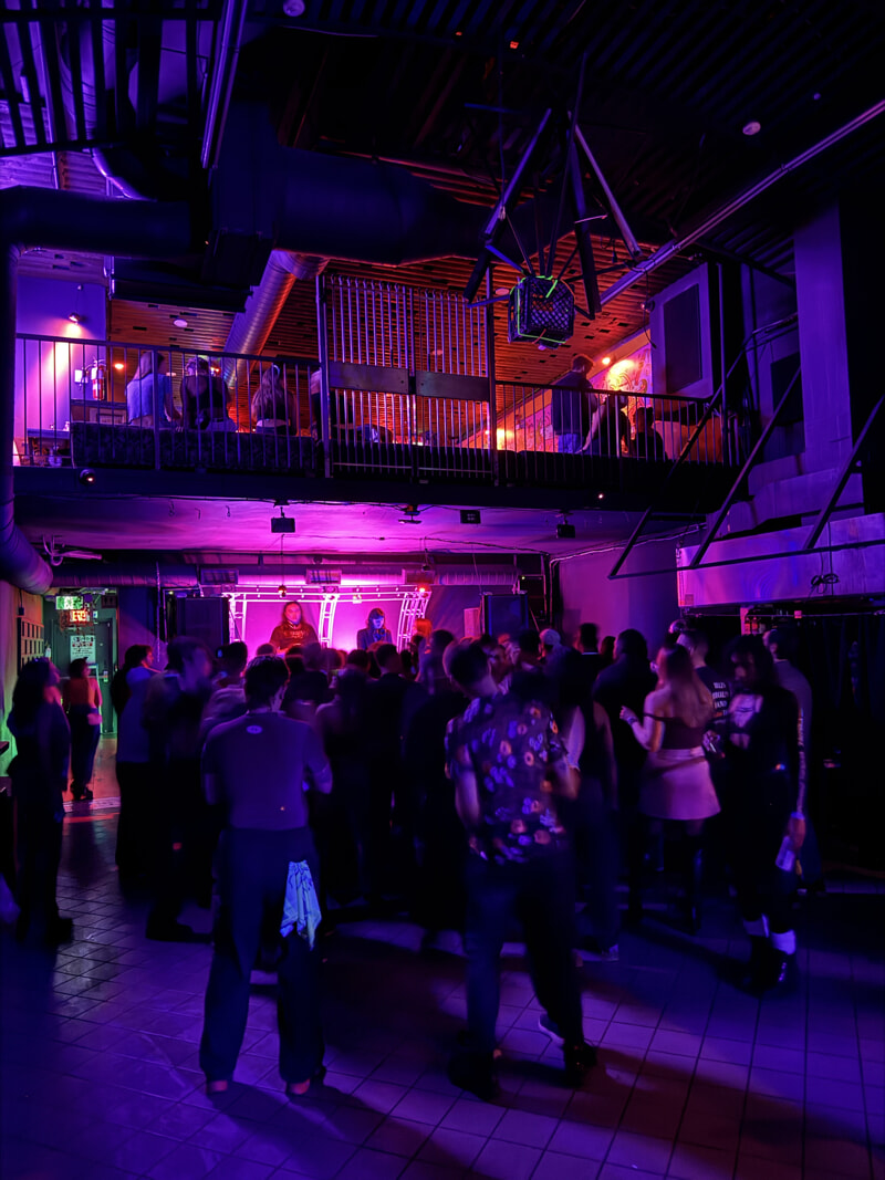 A crowd of people stands looking towards a DJ in an alcove near the centre of the frame.