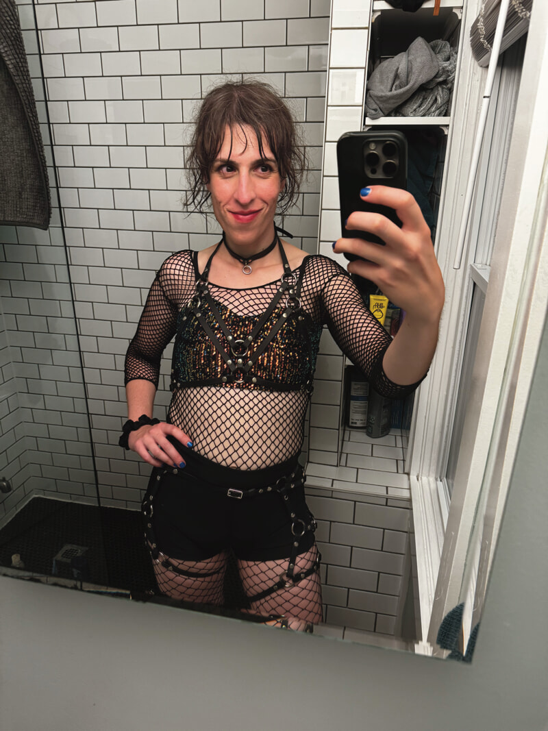 Bathroom mirror selfie. My hair is a mess. I'm wearing a large-mesh top under a sparkly bra-like garment. Over my shorts I have a faux-leather harness. I'm smiling; my lipstick is long gone.