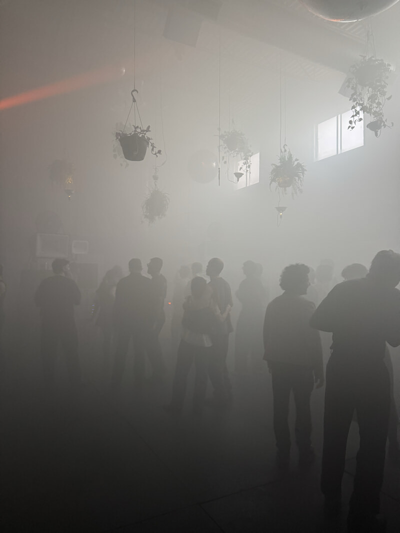 A number of indistinct figures stand in a foggy dance floor. Daylight is coming through the windows.